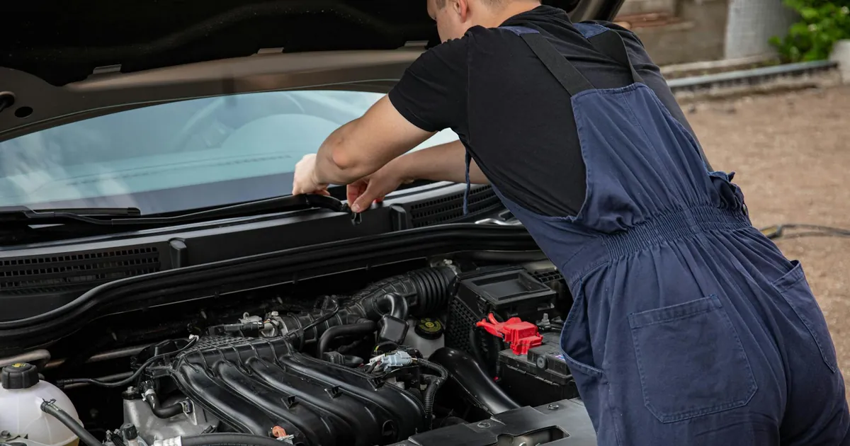 A mechanic in blue overalls works on a car engine outdoors, showcasing vehicle maintenance skills.