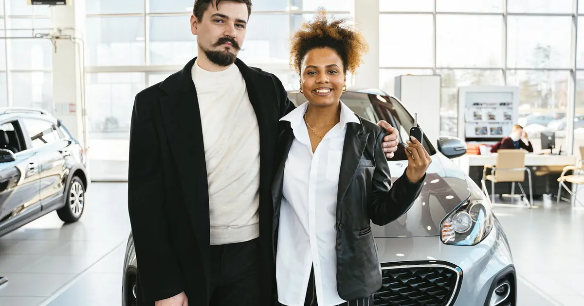 Interracial couple celebrates buying a new car at a dealership, holding keys and smiling.