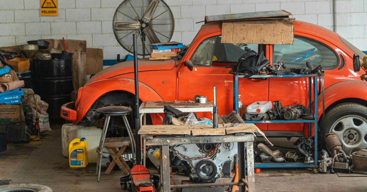 Classic red car being repaired in a cluttered garage setting, filled with tools and auto parts.
