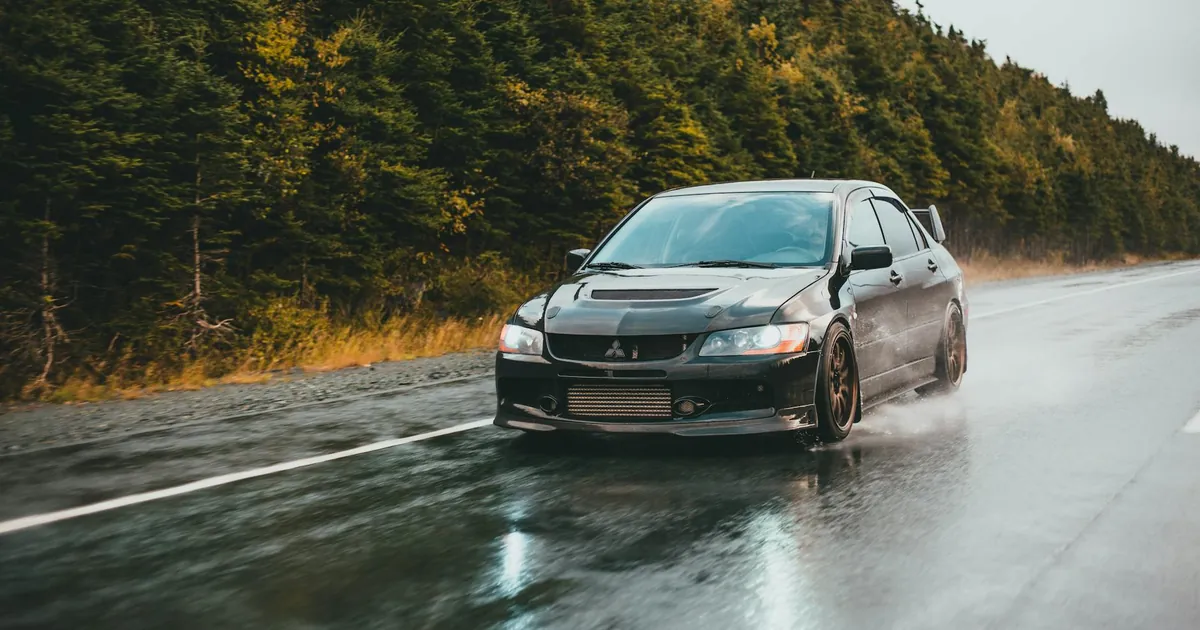 Black Mitsubishi Lancer Evolution speeding down a wet road lined with green trees.