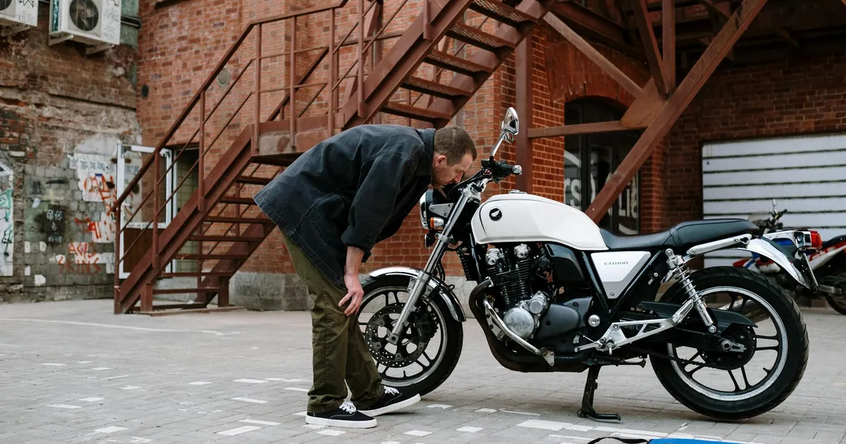 A man inspects a white motorcycle outdoors near a brick building with metal stairs.