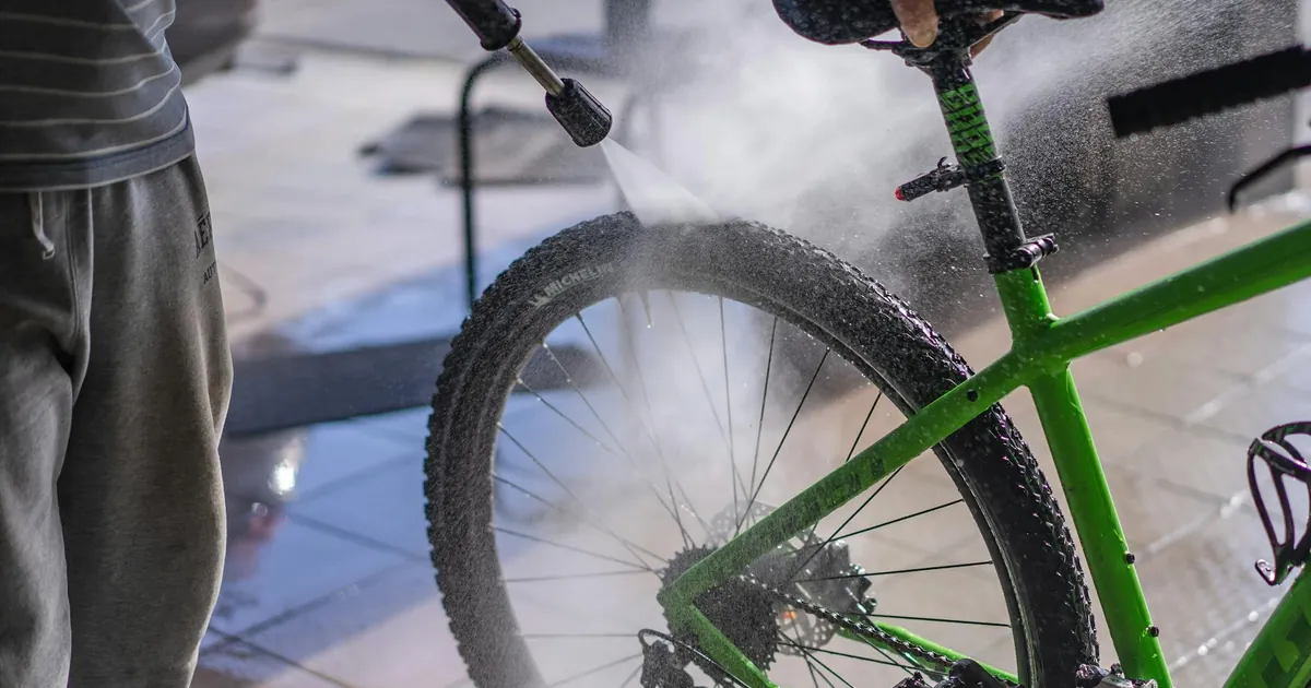 A man cleans a green bicycle with a hose, focusing on the wheel and frame outdoors.
