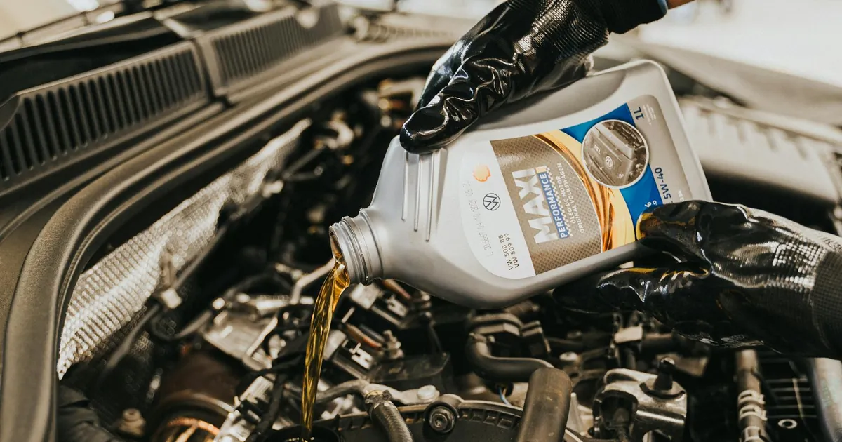 A mechanic wearing gloves pouring oil into a car engine in a garage setting.