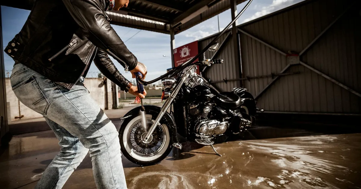 Man cleaning motorcycle with hose at outdoor car wash under sunny skies.