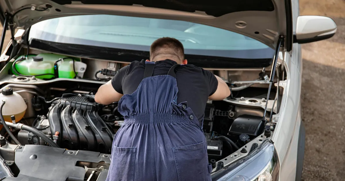 Mechanic in overalls working on a car engine outdoors, offering vehicle repair services.