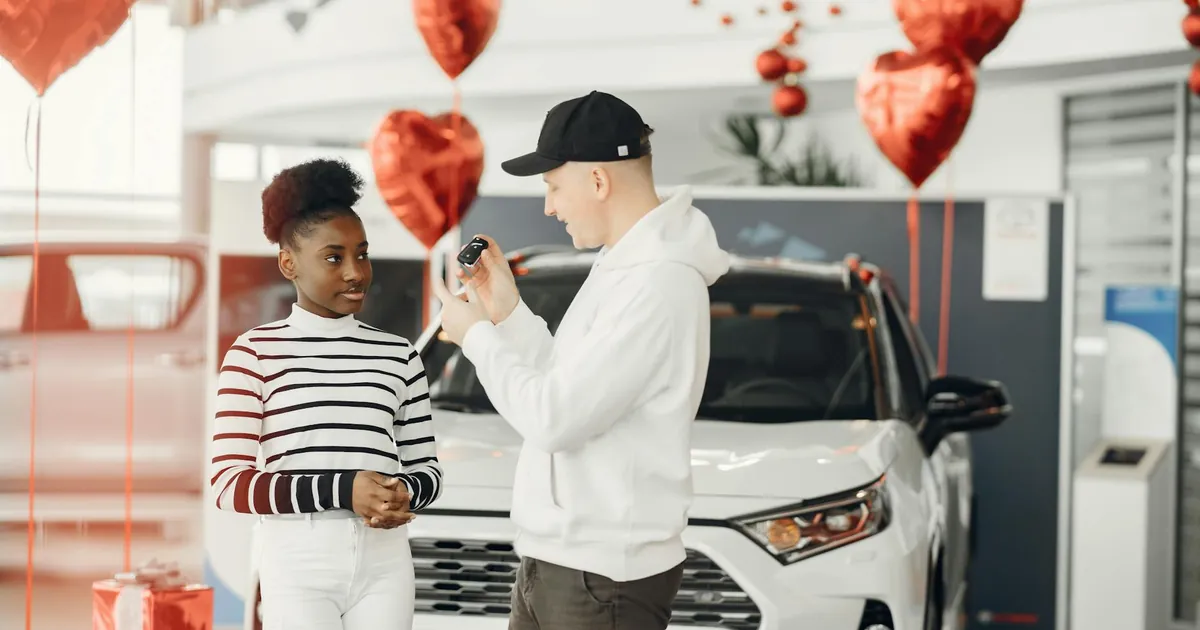 A couple exchanging car keys inside a showroom decorated with heart-shaped balloons, celebrating a new car purchase.