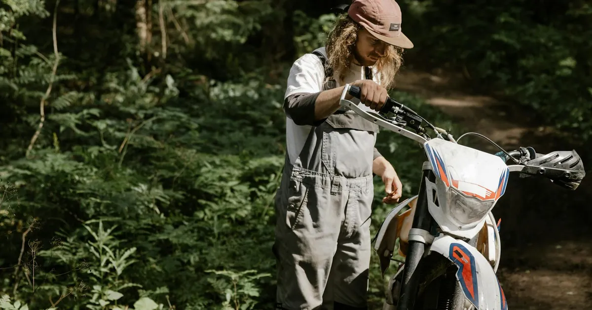 A man in overalls adjusts a dirt bike in a lush forest, highlighting outdoor adventure.