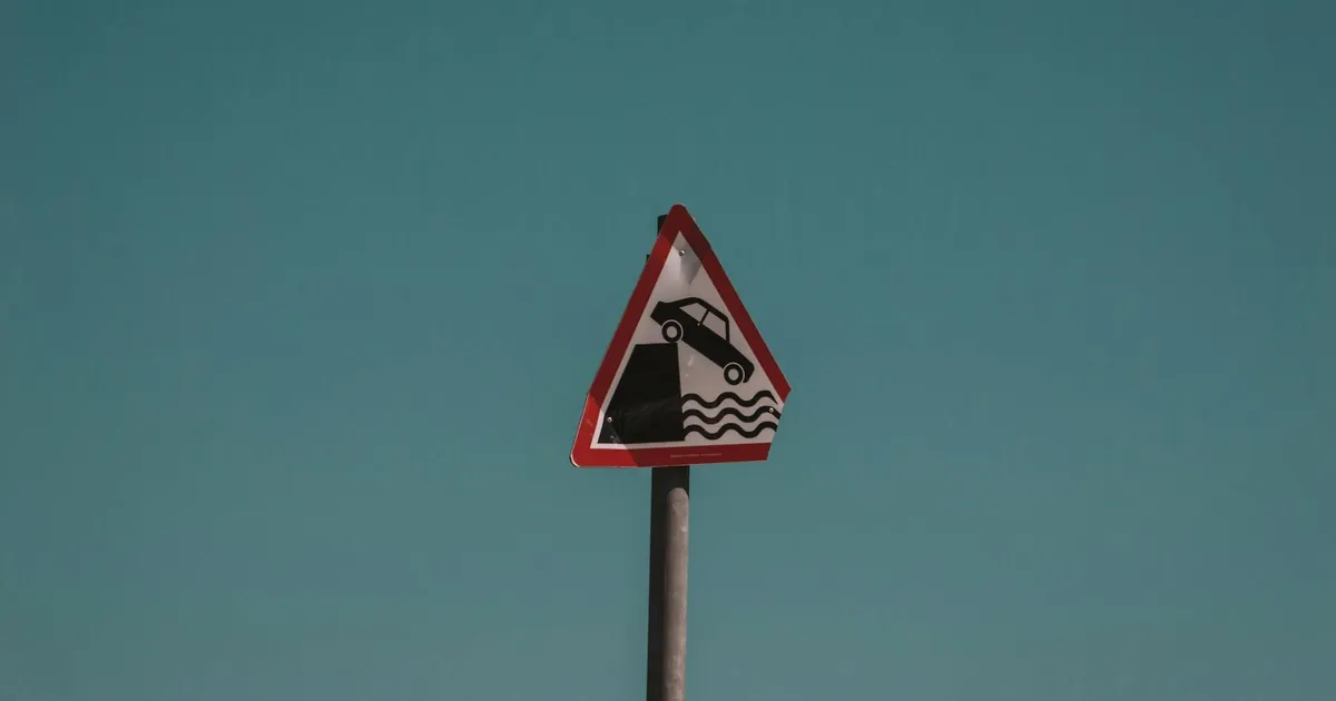 Triangular traffic warning sign showing a car falling into water, set against a clear blue sky in Dublin, Ireland.