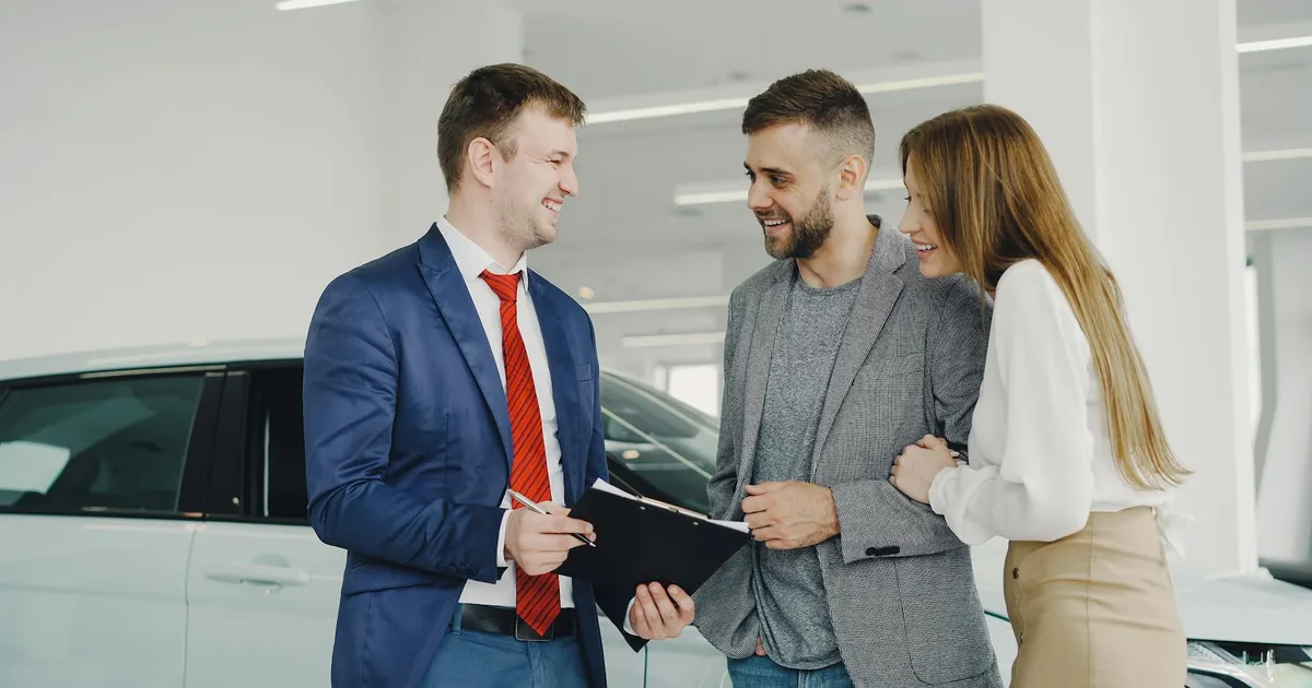 A smiling couple buys a new car from a confident salesman inside a modern car dealership.